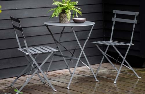 The Hawaii 2-seater bistro set with small round table made from metal and finished in charcoal grey on a stone patio with black horizontal panelled fence