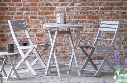 The Miami 2-seater bistro set with small round table made from wood and finished in white on a stone patio with red brick wall